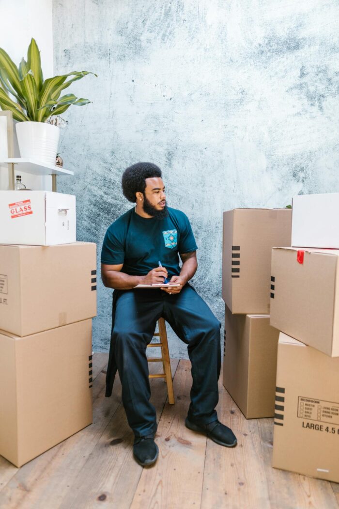 A man sitting on a chair surrounded by cardboard boxes, taking notes.