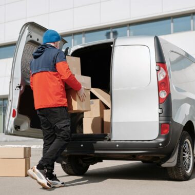 Man in colorful jacket loading cardboard boxes into a van outside an office building.